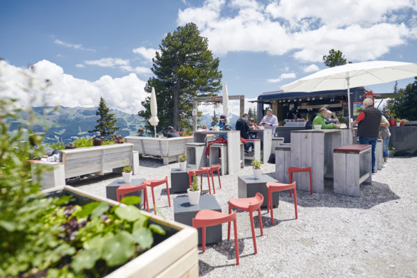 Außenbereich eines Bergrestaurants mit Gästen an Tischen unter Sonnenschirmen, umgeben von grünen Pflanzen und mit Berglandschaft im Hintergrund bei sonnigem Wetter.